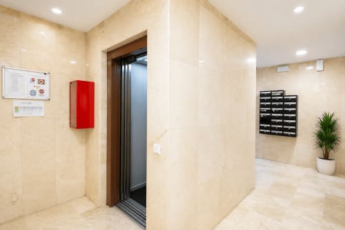 Vestíbulo moderno con paredes de mármol beige, puertas de ascensor abiertas, extintor rojo y buzones en Riurau Home