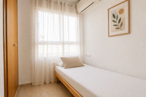 Luminoso dormitorio individual en apartamento Riurau Home de Calpe con cama de madera, cortinas blancas, aire acondicionado y decoración minimalista