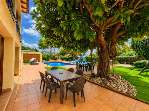 Casa Emilia villa terrace with dining table under leafy tree, blue pool and gardens at Moraira