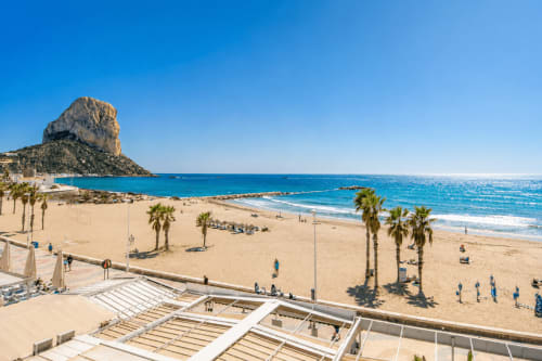 Calpe beach with golden sand, palm trees, turquoise Mediterranean Sea, and iconic Peñón d'Ifach rock formation under clear