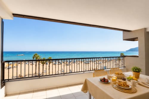 Balcón de apartamento frente al mar en Calpe con vistas al océano, mesa de comedor y playa arenosa con palmeras