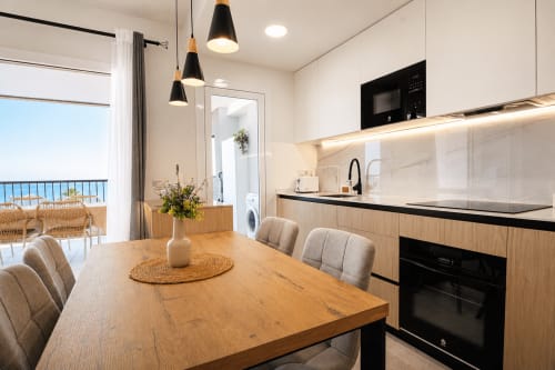 Cocina y comedor con vistas al mar en el apartamento Riurau Home Calpe, con mesa de madera, luminarias colgantes negras y estilo costero