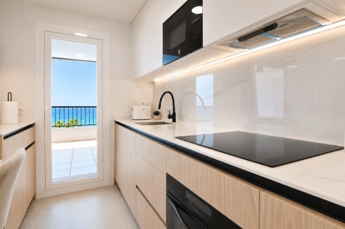 Cocina moderna con vistas al mar en apartamento de Calpe, con muebles de madera clara, encimera negra y balcón con vista