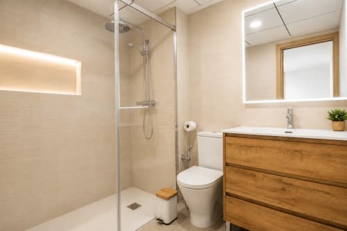 Baño moderno en apartamento Riurau Home Calpe con ducha abierta, lavamanos de madera, espejo iluminado y accesorios blancos