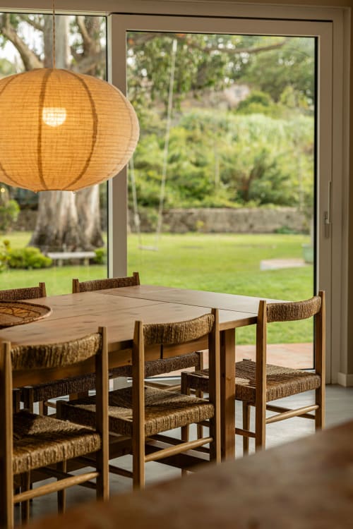 Dining area with wooden table, woven chairs, and orange pendant lamp overlooking garden at Sea'Ya Casa Pavão villa in Colares