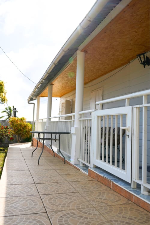 Patio du gîte R'marine439 à Sainte-Rose avec terrasse carrelée, balustrades blanches, véranda couverte et vue sur le jardin