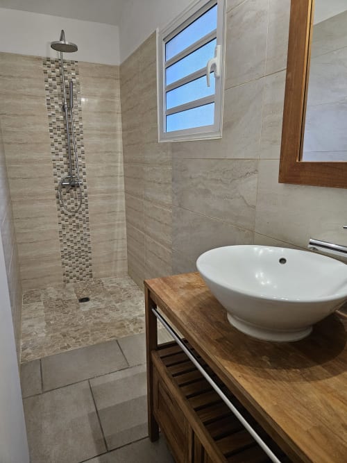 Salle de bain moderne à la Villa Cœur des laves (R'marine439) avec douche pluie, lavabo blanc rond, meuble en bois et vue sur mer