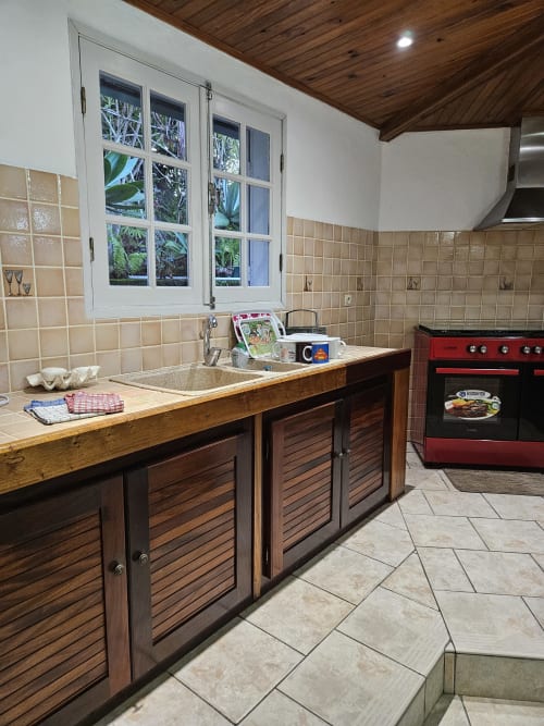 Cuisine de villa moderne avec armoires en bois, dosseret en carrelage beige, cuisinière rouge et fenêtre blanche surplombant les jardins tropicaux