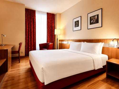 Chambre élégante de l'Hôtel De La Poste à Esch sur Alzette avec lit queen-size, mobilier en bois, rideaux rouges et vue sur la ville