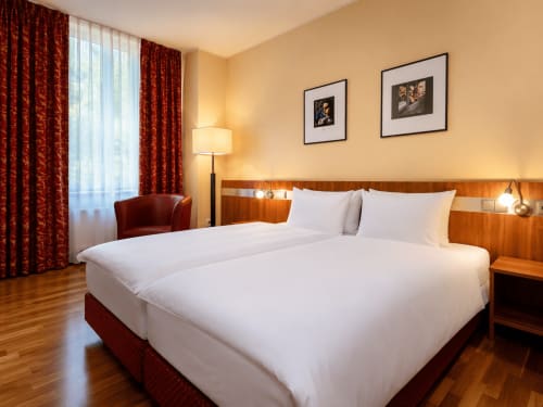 Chambre d'hôtel moderne à l'Hôtel De La Poste à Esch sur Alzette avec lits jumeaux, mobilier en bois, rideaux rouges et vue sur la ville