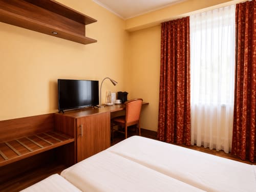 Chambre confortable à l'Hôtel De La Poste à Esch-sur-Alzette avec bureau en bois, TV et vue sur la ville par la fenêtre aux volets rouges