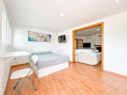Bright villa bedroom in Reus with wooden flooring, queen bed, folding chair, wall-mounted TV, and doorway to living area with