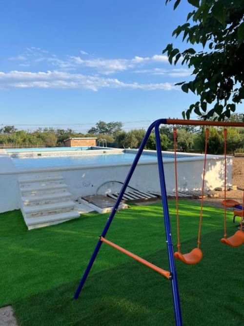 Blue and orange swing set on artificial grass beside outdoor swimming pool at Rentalaya villa in Reus with trees and clear