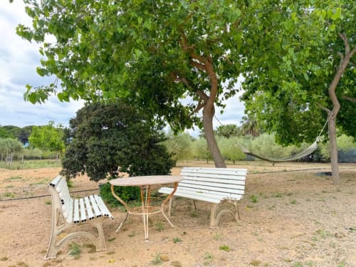 White metal outdoor furniture and hammock under shaded trees in the garden area of Rentalaya villa in Reus, Spain