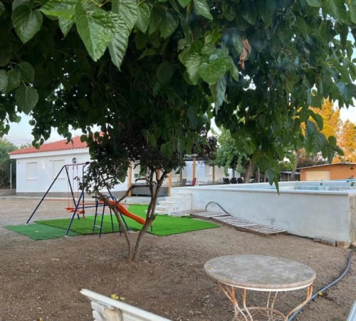 Garden area of Rentalaya villa in Reus with playground, large tree, outdoor furniture, and swimming pool