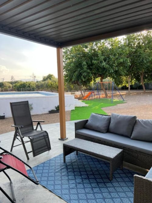 Covered patio with gray outdoor seating, lounger chair, and blue rug overlooking private pool and garden at Rentalaya villa