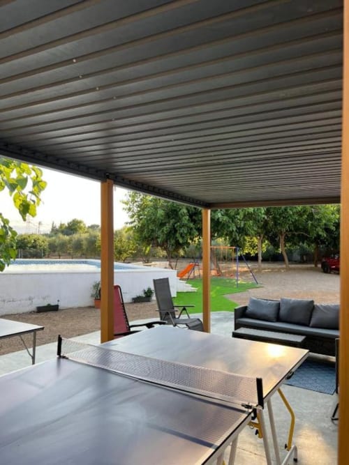 Covered patio area at Rentalaya villa in Reus with ping-pong table, black sofa, lounge chairs, and garden views