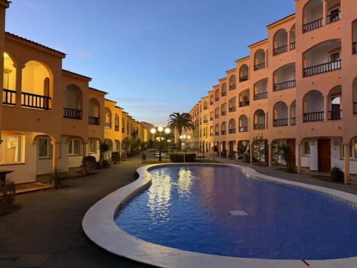 Peñíscola resort courtyard at dusk with curved pool, terracotta apartment buildings, palm trees, and ambient lighting