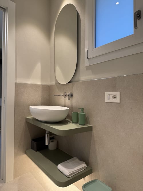 Bagno moderno con specchio ovale, lavabo bianco sospeso e dettagli verdi nell'appartamento La Casetta dei Ronchi a Marina di Massa