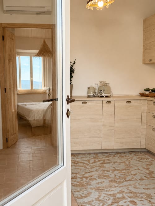 Tuscan Townhouse kitchen with wooden cabinetry, sea view through open bedroom door, beige stone counters and patterned rug