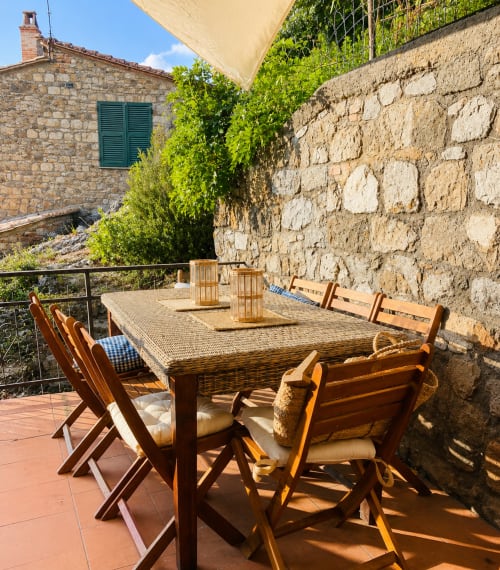 Tuscan stone terrace with wooden dining table and chairs, shaded by beige umbrella, overlooking gardens at Montemassi