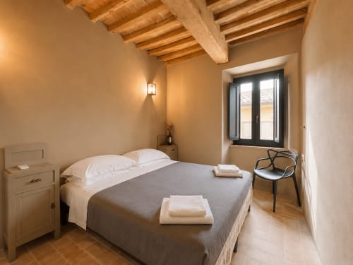 Tuscan townhouse bedroom with exposed wooden beams, terracotta floor, white bedding, and black-framed windows in Montemassi,