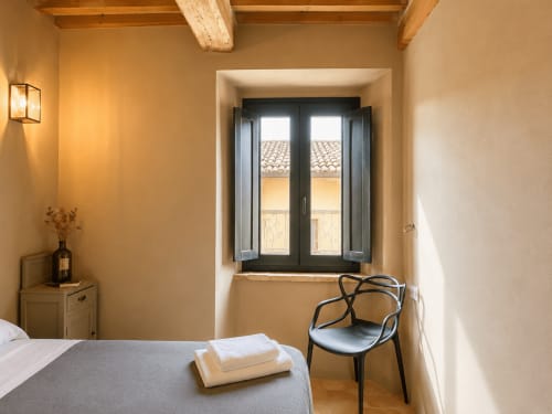 Bright Tuscan bedroom with double bed, black modern chair, wooden beams, and dark double doors opening to countryside views