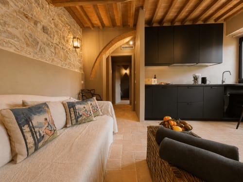Tuscan townhouse apartment with stone walls, wooden beams, cream sofa, black kitchen, and terracotta floors in Montemassi