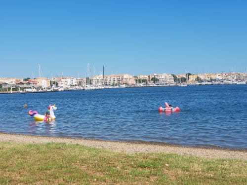 Enfants jouant dans l'eau peu profonde avec des bouées gonflables à la plage de Cap d'Agde, marina et voiliers en arrière-plan