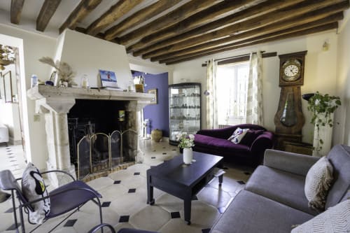 Élégant salon du Domaine Chapelle avec cheminée, poutres apparentes, canapé violet et horloge de parquet ancienne