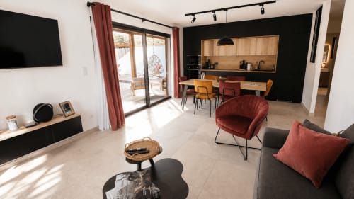 Spacieuse zone de vie d'une villa moderne en Corse avec cuisine, table à manger, fauteuil rouge, canapé, TV et portes vitrées donnant sur la terrasse