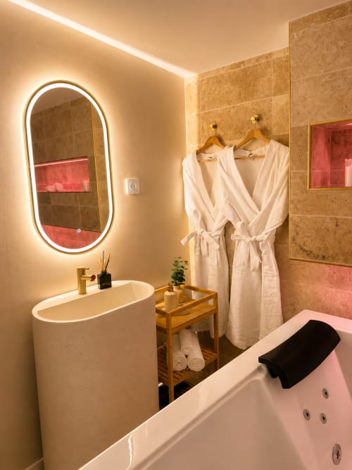 Salle de bain moderne à l'hôtel L'Escapade au Mans avec miroir ovale illuminé, vasque en pierre, baignoire et peignoirs blancs
