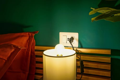Calaba Decumani bedroom detail: teal wall, wooden headboard, red bedding, white lamp, and wall outlet in Naples