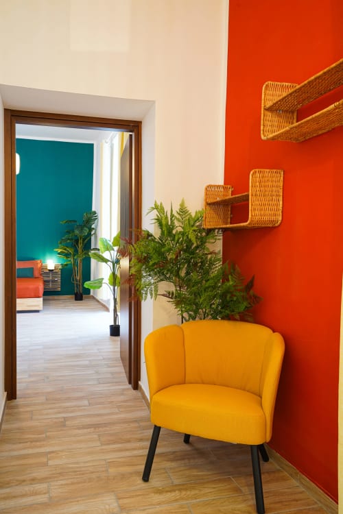 Colorful Calaba apartment entrance in Naples featuring yellow chair, red wall, wicker shelves, and view to teal bedroom