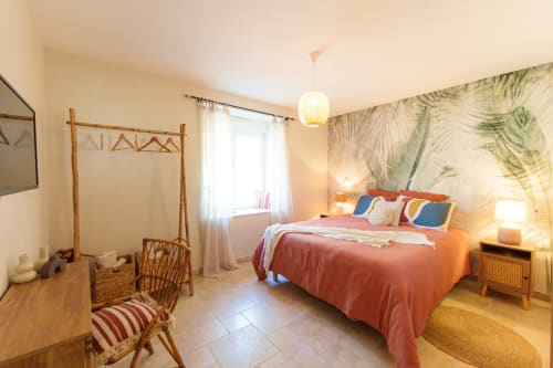 Chambre lumineuse au Hameau de Céline avec couvre-lit corail, lit à baldaquin en bambou, papier peint tropical et vue sur le jardin.