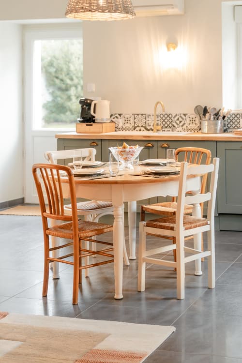 Salle à manger lumineuse avec table ronde en bois, chaises mixtes bois et blanc, suspension en rotin, et cuisine moderne au Hameau