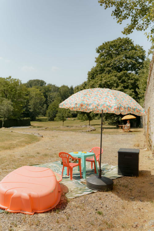 Zone de détente colorée au Hameau de Céline avec parasol orange, table turquoise et chaises longues dans le jardin des Landes