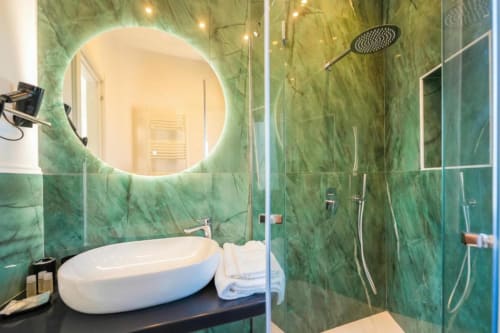 Bagno moderno presso Origenes Rooms Procida con pareti in marmo verde, specchio circolare illuminato, lavabo ovale bianco e doccia in vetro