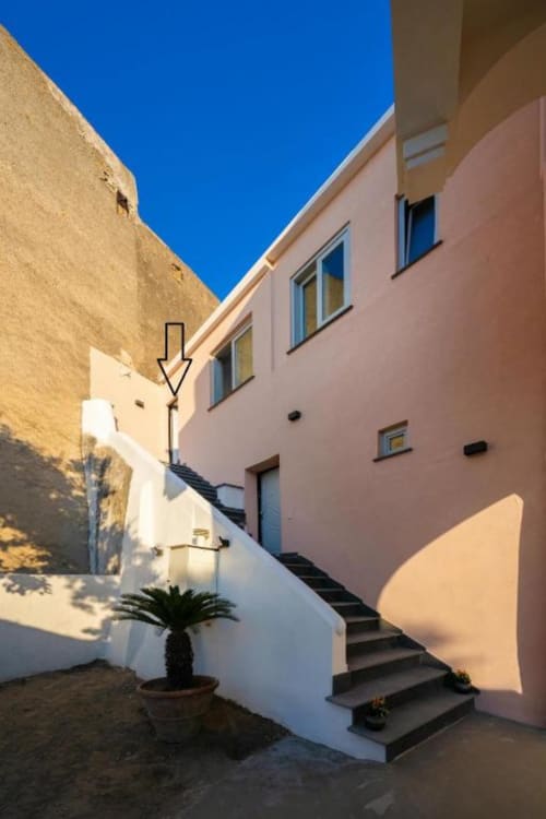 Affascinante cortile mediterraneo di Origenes Rooms a Procida con edifici bianchi e rosa, scale in pietra e cielo azzurro