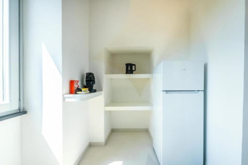 Moderna kitchenette con mobili bianchi, ripiani aperti, frigorifero e bollitore elettrico nell'appartamento Ariè di Origenes