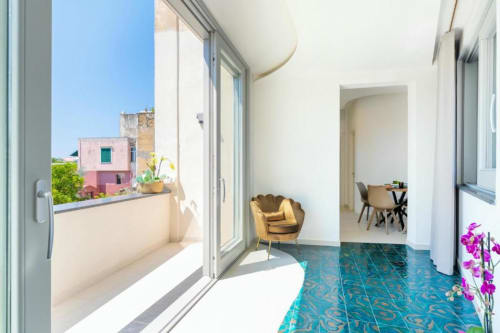 Luminoso appartamento moderno presso Origenes Rooms a Procida con pavimento in piastrelle blu, sedia dorata, porta sulla terrazza e edificio colorato