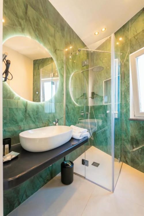 Moderno bagno dell'appartamento Ariè presso Origenes Rooms, Procida, con piastrelle in marmo verde, specchio circolare illuminato e lavabo bianco