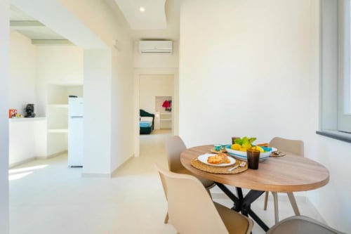 Zona pranzo moderna nell'appartamento Ariè di Origenes Rooms, Procida, con tavolo in legno, sedie beige e vista della cucina