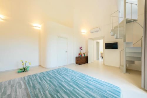 Camera moderna nell'appartamento Ariè presso Origones Rooms, Procida, con letto a righe blu, scala a spirale e luminosa planimetria aperta.
