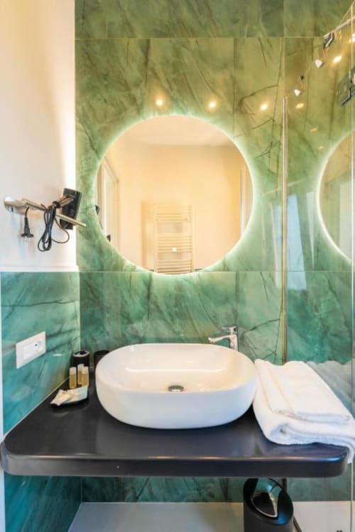 Moderno bagno di Origenes Rooms, Procida: pareti in marmo verde, specchio circolare illuminato, lavabo bianco su nero