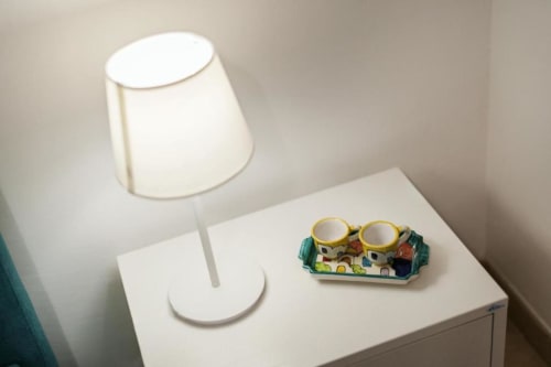 Lampada da tavolo bianca con portabicchieri decorativo con casette verdi su comodino presso l'appartamento Origenes Rooms