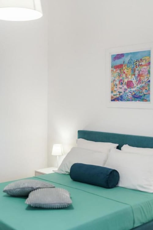 Camera da letto moderna nell'appartamento di Procida con letto turchese, cuscini bianchi e coloratissime opere d'arte costiera
