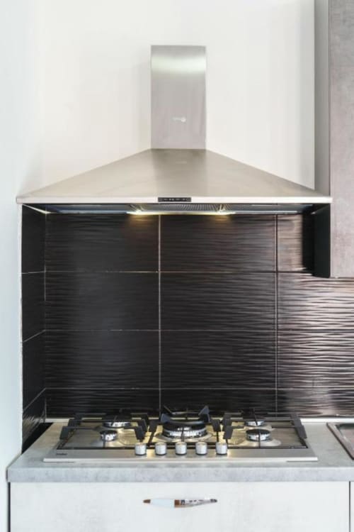 Cucina moderna con piano cottura a gas, cappa aspirante in acciaio inox e piastrelle nere texture in appartamento Origenes Rooms,