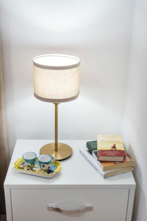 Comodino in Origenes Rooms con lampada color crema, pila di libri e tazze decorative sul comodino bianco