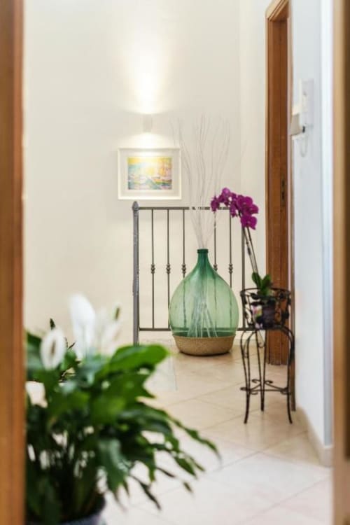 Corridoio luminoso alle Origenes Rooms, Procida, con piante decorative, vaso verde e ringhiera metallica che conduce alla terrazza